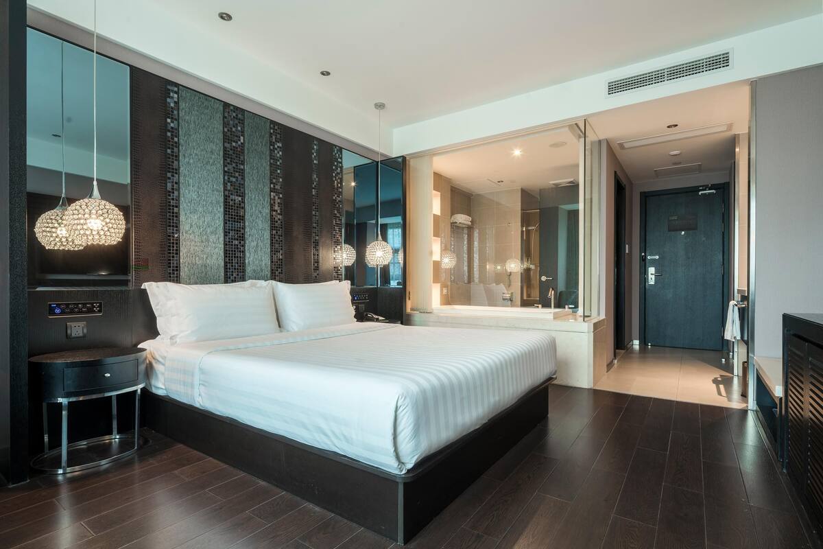 Ein modernes Hotelzimmer mit einem großen Bett, Bedienelementen am Bett, einem runden Nachttisch und einem großen Spiegel über dem Bett.