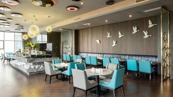 Un interior de restaurante moderno con mesas redondas, sillas color turquesa y una pared decorada con motivos de pájaros.