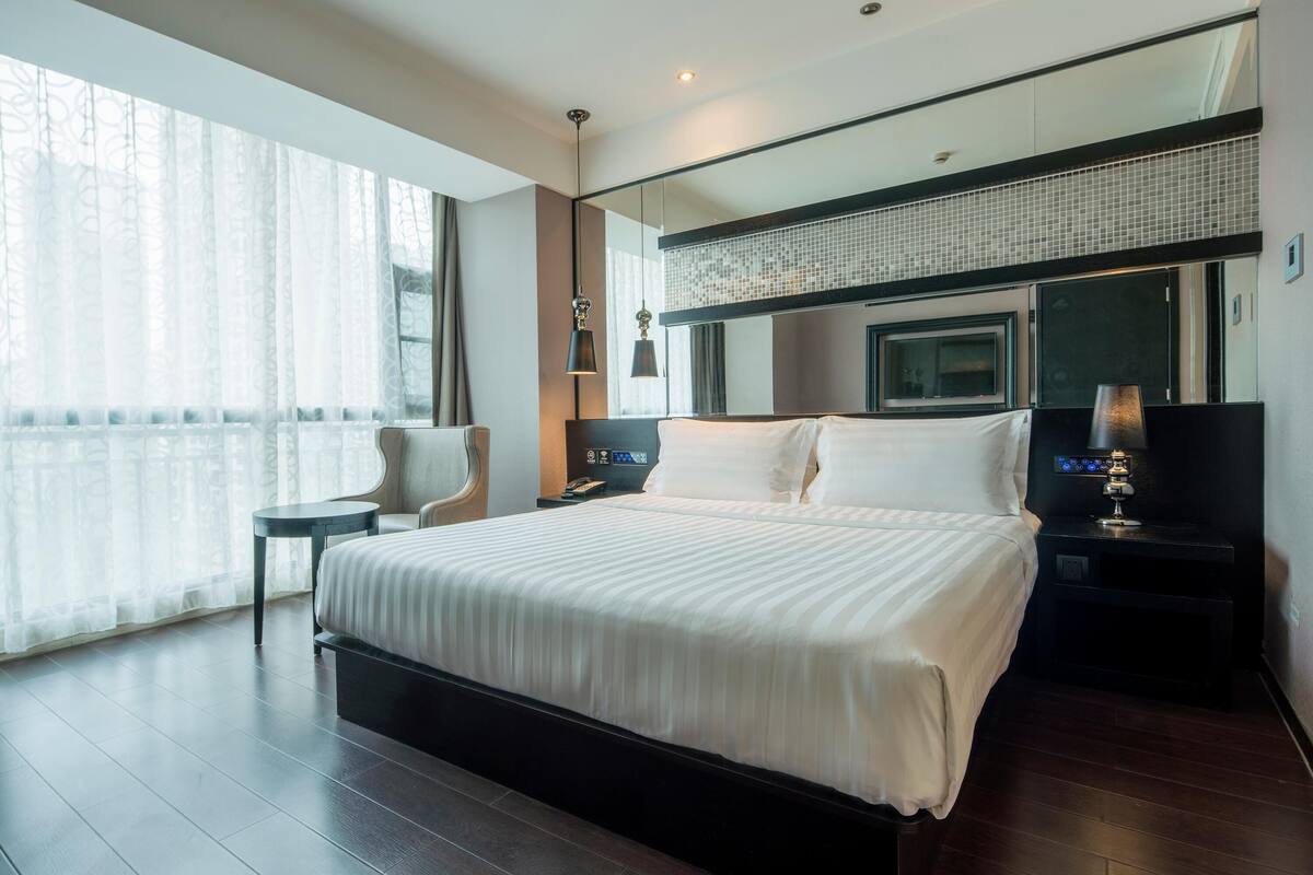 Ein modernes Hotelzimmer mit einem großen Bett, einem Nachttisch mit Lampe, einem kleinen Beistelltisch und einem großen Fenster mit durchsichtigen Vorhängen.