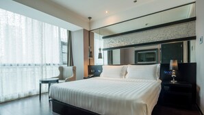 A modern hotel room with a large bed, a bedside table with a lamp, a small side table, and a large window with sheer curtains.