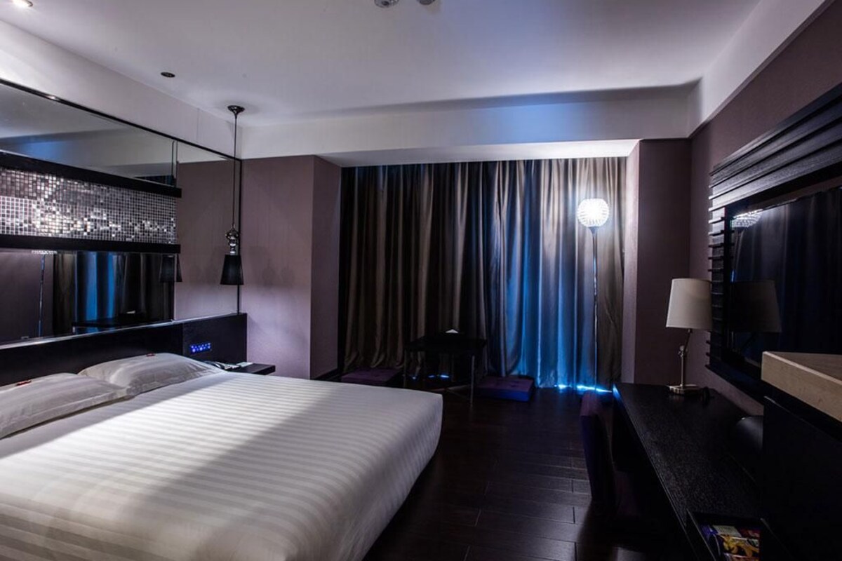 A modern hotel room with a large bed, a desk, a mirror, and a view of the city through curtains.