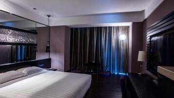 A modern hotel room with a large bed, a desk, a mirror, and a view of the city through curtains.