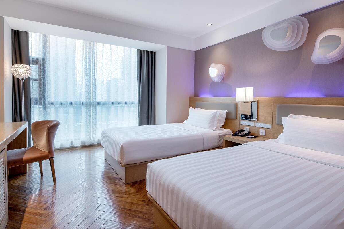 Ein Hotelzimmer mit zwei Betten, einem Schreibtisch, einem Stuhl und einem großen Fenster mit Vorhängen.