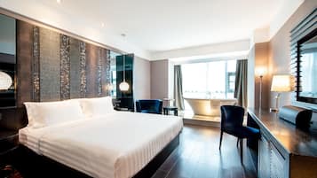 A modern hotel room with a large bed, a desk, a chair, a small table, and a view of the city through large windows.