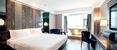 Una habitación de hotel moderna con una cama grande, un escritorio, una silla, una mesita y vistas a la ciudad a través de amplios ventanales.