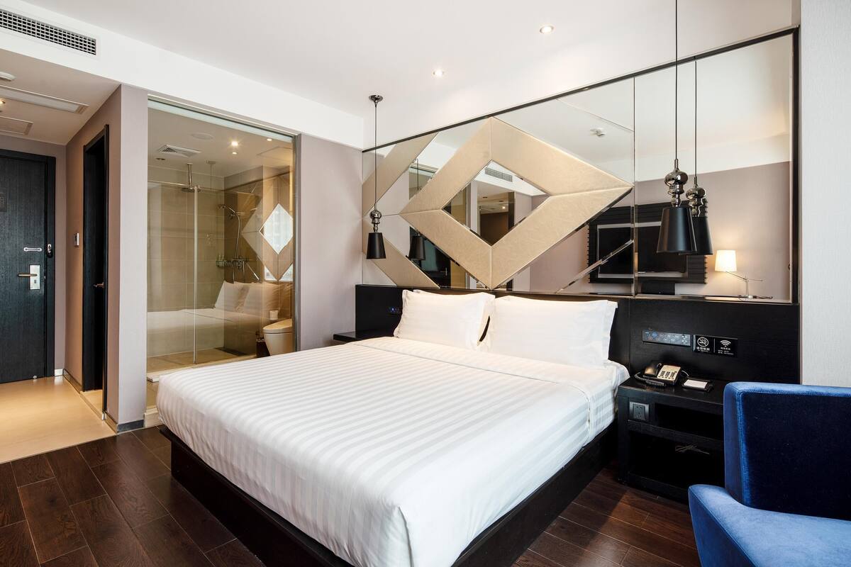 Ein modernes Hotelzimmer mit einem großen Bett, einem Schreibtisch mit Lampe und einem Spiegel mit geometrischen Mustern.