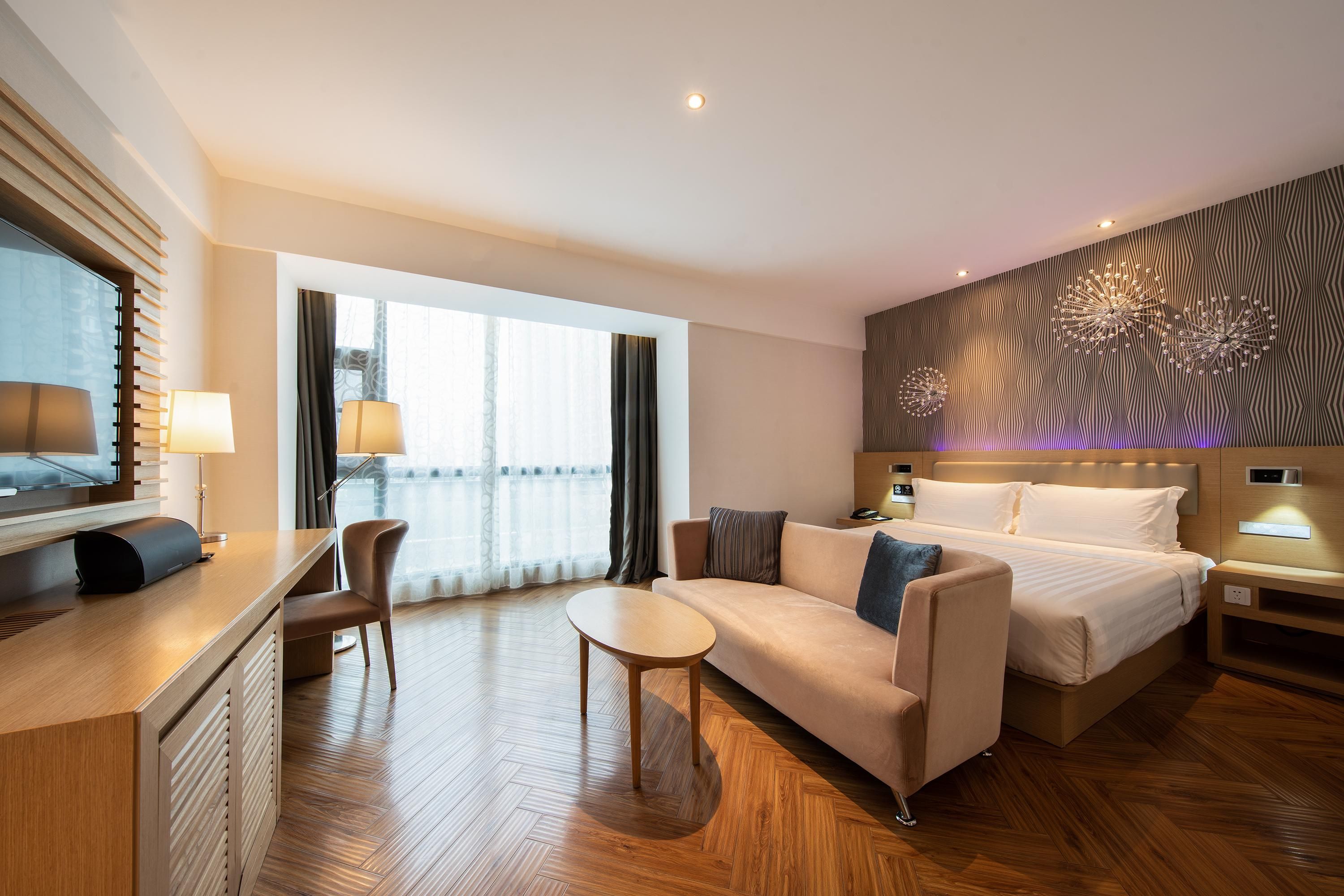 A modern hotel room with a bed, sofa, desk, and a large window.