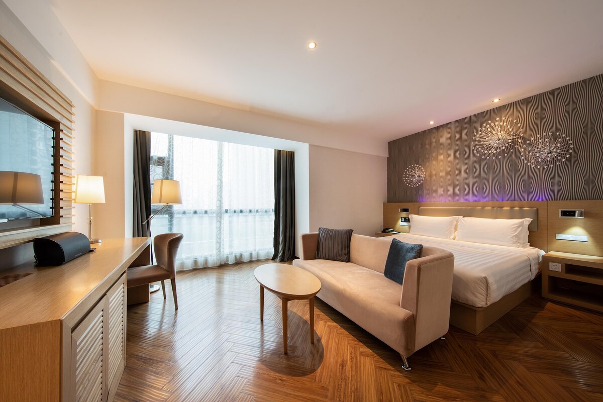 A modern hotel room with a bed, sofa, desk, and a large window.