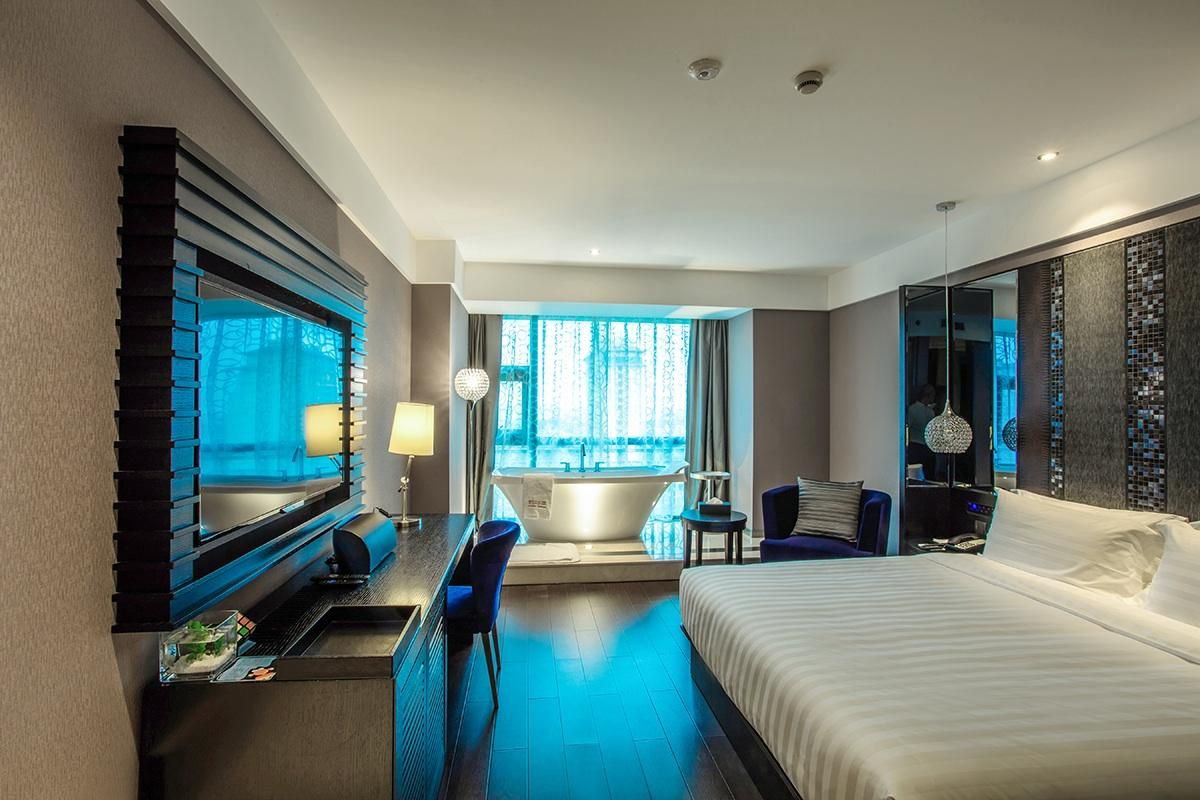A modern hotel room with a large bed, a desk with a chair, a bathtub, and a large window with curtains.