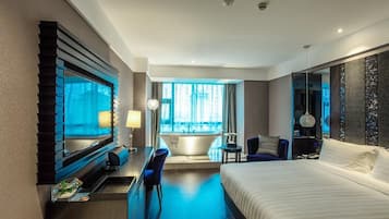 A modern hotel room with a large bed, a desk with a chair, a bathtub, and a large window with curtains.