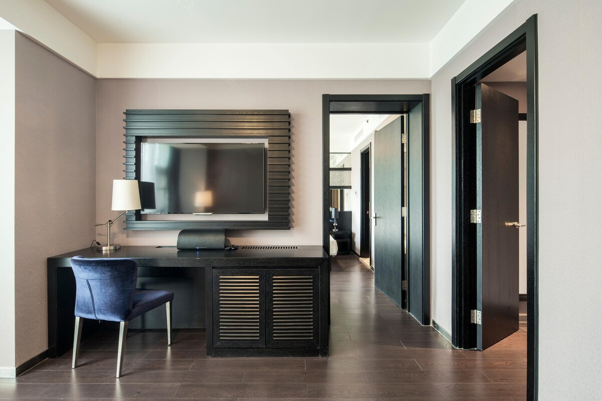 Une chambre d’hôtel moderne avec un bureau en bois foncé, une chaise bleue, un grand téléviseur à écran plat fixé au mur et une porte en bois foncé.