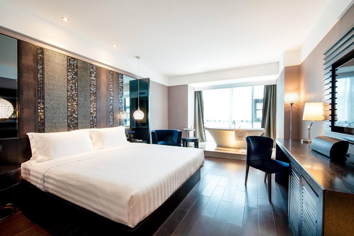 Ein modernes Hotelzimmer mit einem großen Bett, einem Schreibtisch, einem Stuhl, einem kleinen Tisch und Blick auf die Stadt durch große Fenster.