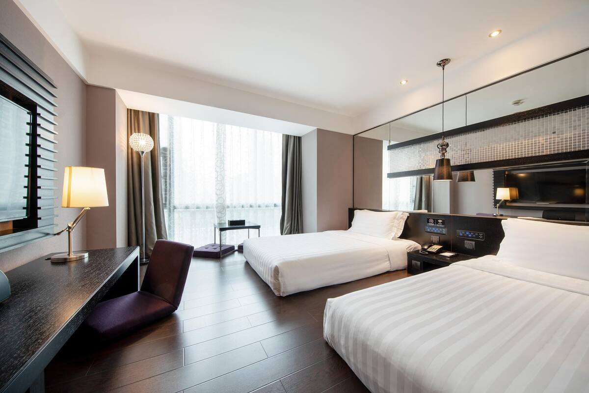 Ein Hotelzimmer mit zwei Betten, einem Schreibtisch mit Lampe, einem Sessel, einem Fernseher und einem großen Fenster mit Vorhängen.