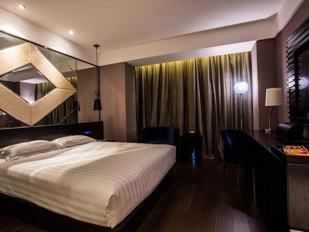 Une chambre d’hôtel moderne avec un grand lit, un bureau et une chaise. Un miroir est accroché au mur et une fenêtre est recouverte de rideaux.