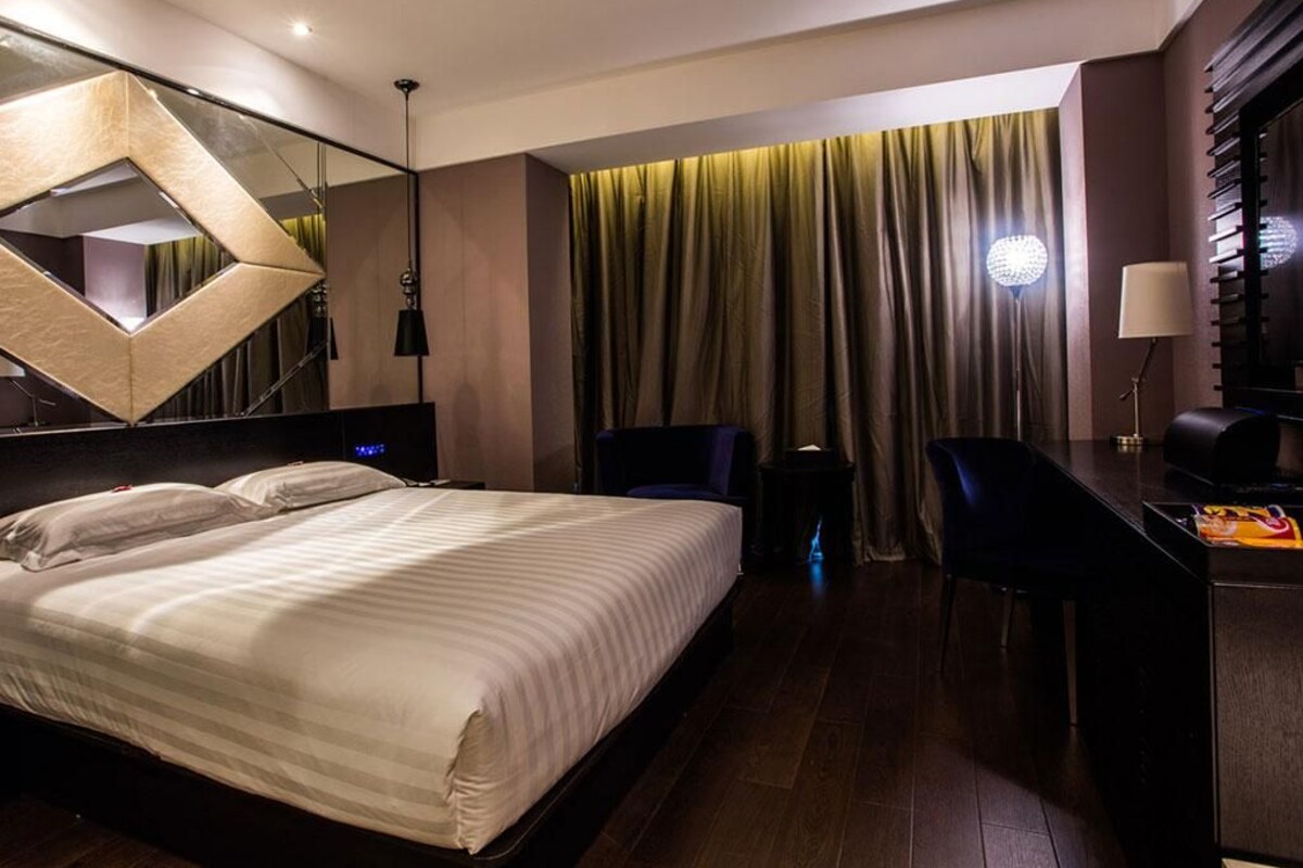 A modern hotel room with a large bed, a desk, and a chair. There is a mirror on the wall and a curtain-covered window.