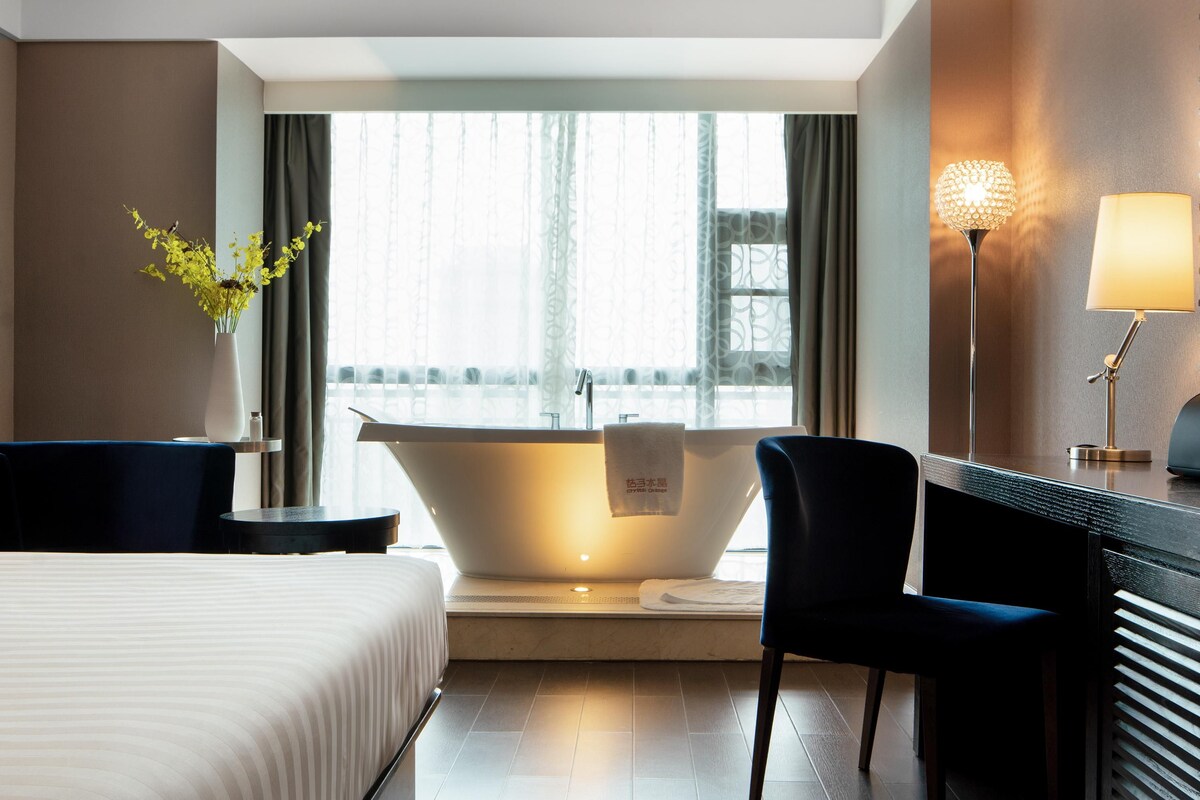Une chambre d’hôtel moderne dotée d’une grande baignoire, d’un lit, d’un bureau avec une lampe et offrant une vue sur la ville grâce à de grandes fenêtres.