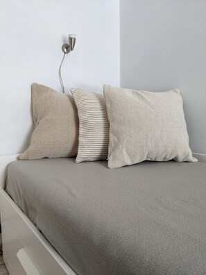 Una cama con un cubrecolcha gris y tres almohadas de diferentes texturas.