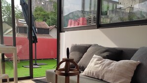 Ein gemütliches Wohnzimmer mit einem grauen Sofa, einem Holztisch mit Obstschale und Gebäck, und einem großen Fenster mit Blick auf einen Innenhof mit roter Mauer und einer Flagge.
