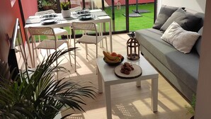 Ein modernes Wohnzimmer mit Glaswand, einem Essbereich mit Tisch für Zwei, einem grauen Sofa und einem kleinen Garten, der durch das Glas sichtbar ist.