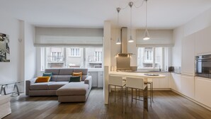 Un apartamento moderno con sala de estar que incluye un sofá y una mesa de comedor con sillas, una cocina con estufa y horno, y amplias ventanas con balcón.