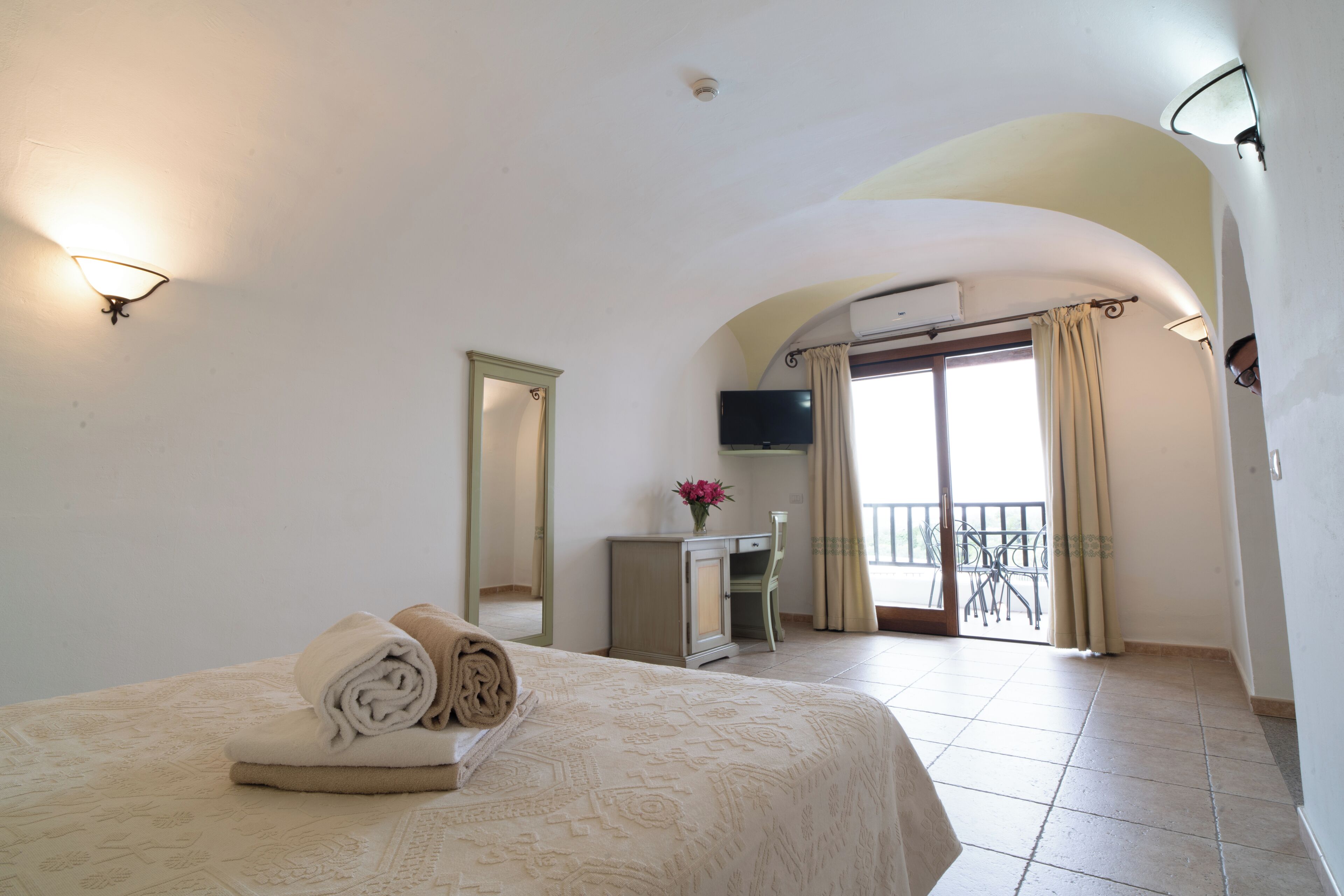 A hotel room with a bed, a mirror, a TV, a desk, and a balcony with a view.