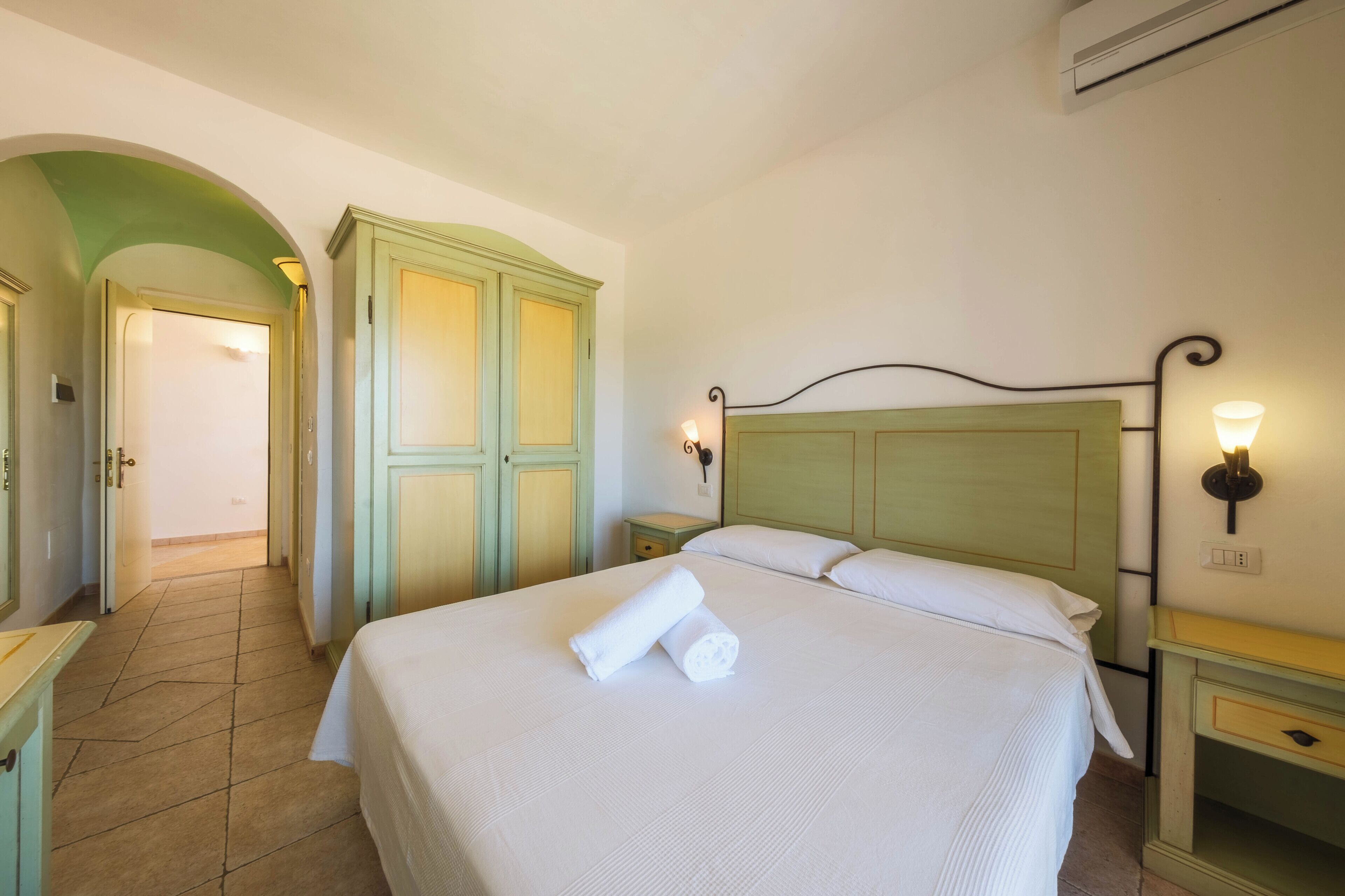 Una habitación de hotel con cama, mesita de noche, lámpara, armario y un arco de entrada.