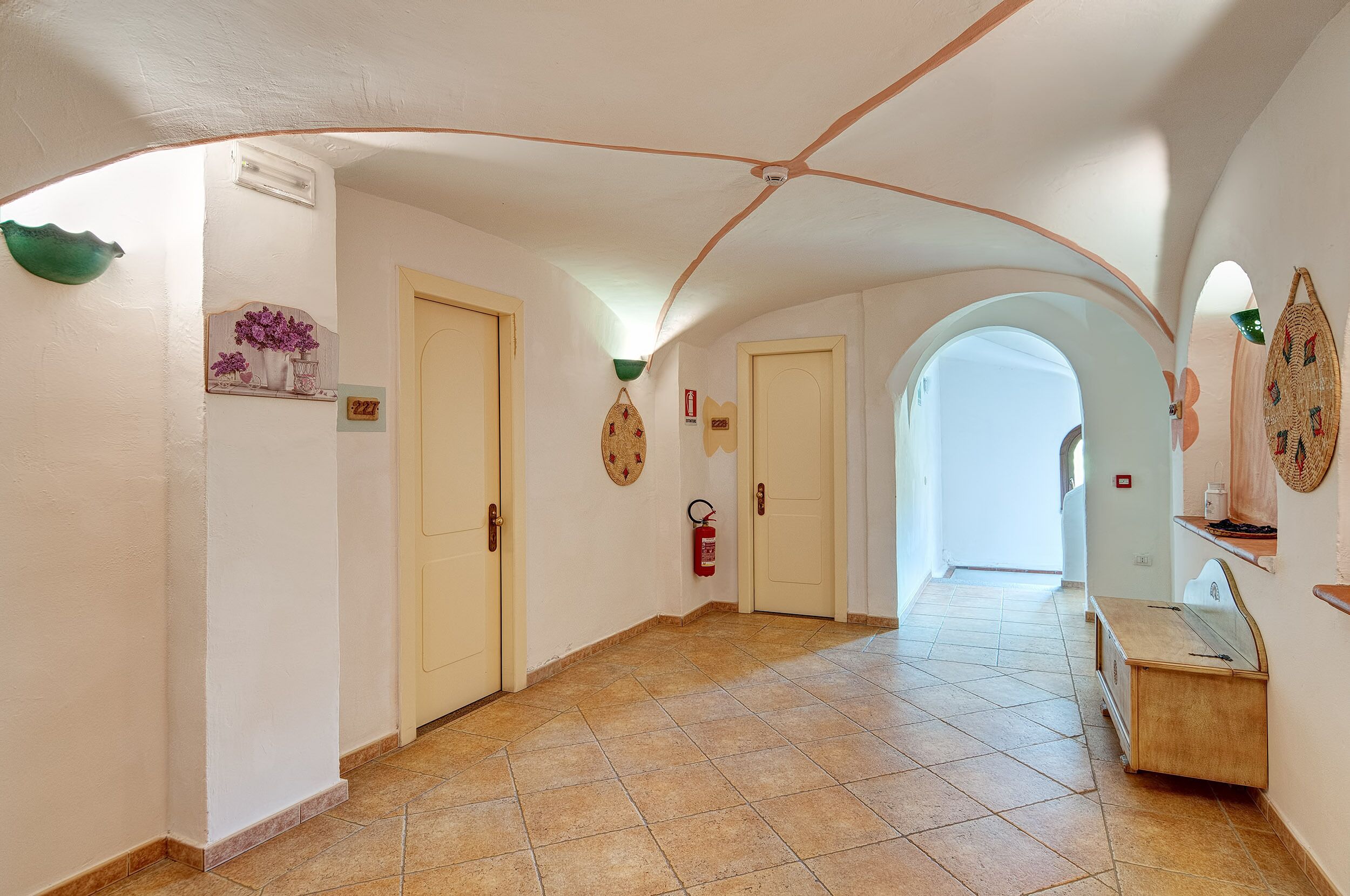 A hallway with white walls, a tiled floor, and a ceiling with a curved design. There are doors, a fire extinguisher, and a small table with a vase of flowers.