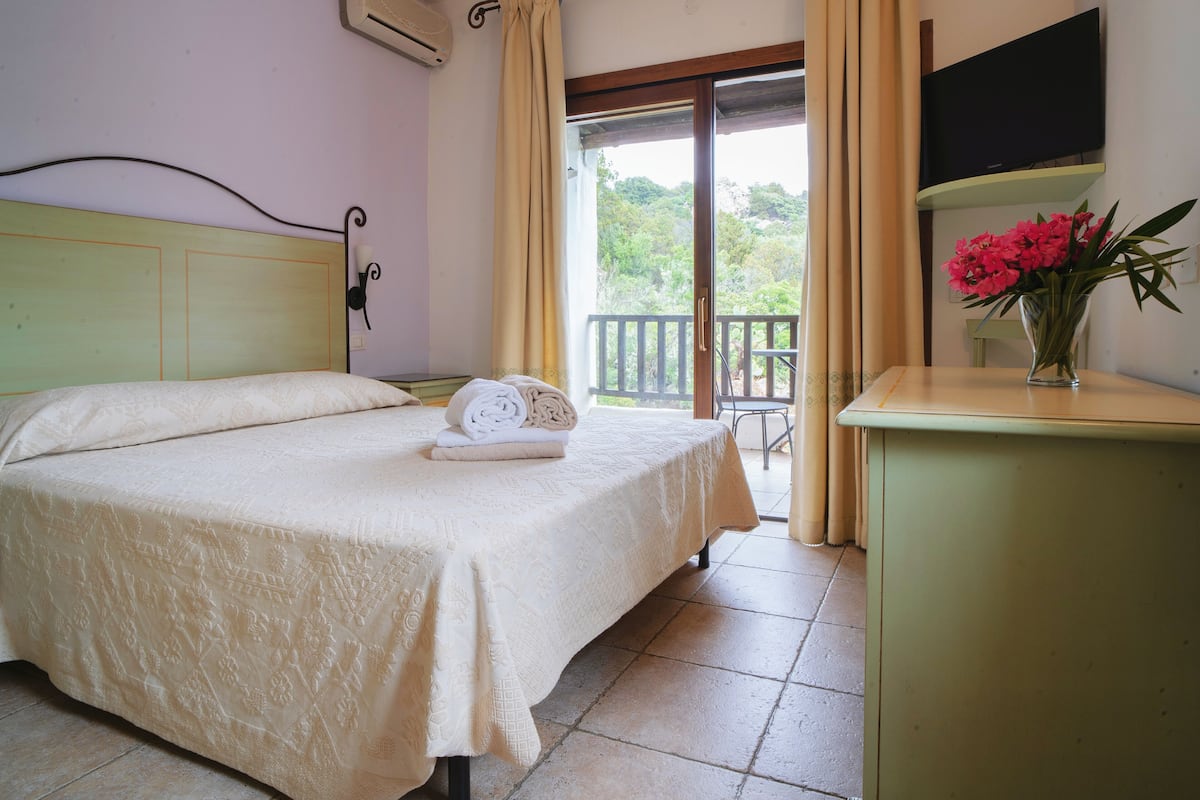 A bedroom with a bed, a bedside table with a vase of flowers, a TV mounted on the wall, and a balcony with a view of greenery.