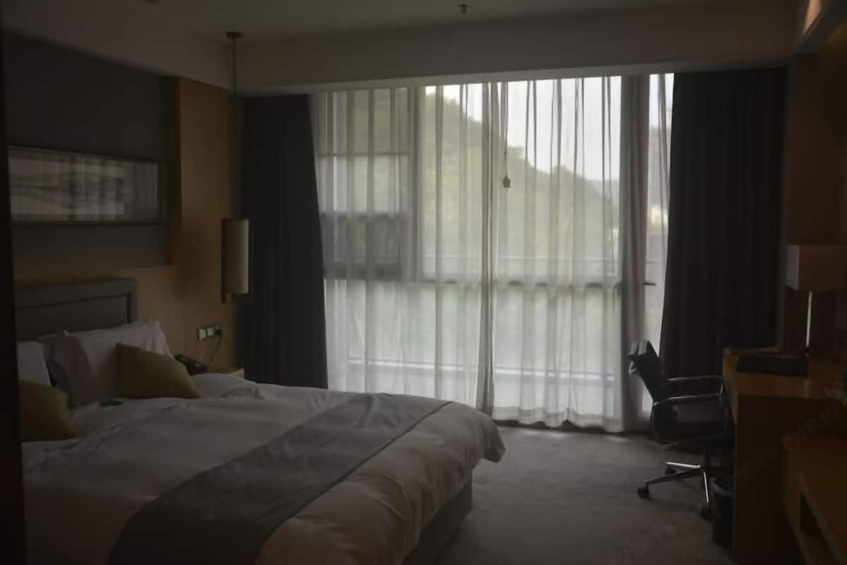 A hotel room with a large bed, a desk, and a chair. There is a window with curtains, and a painting on the wall.