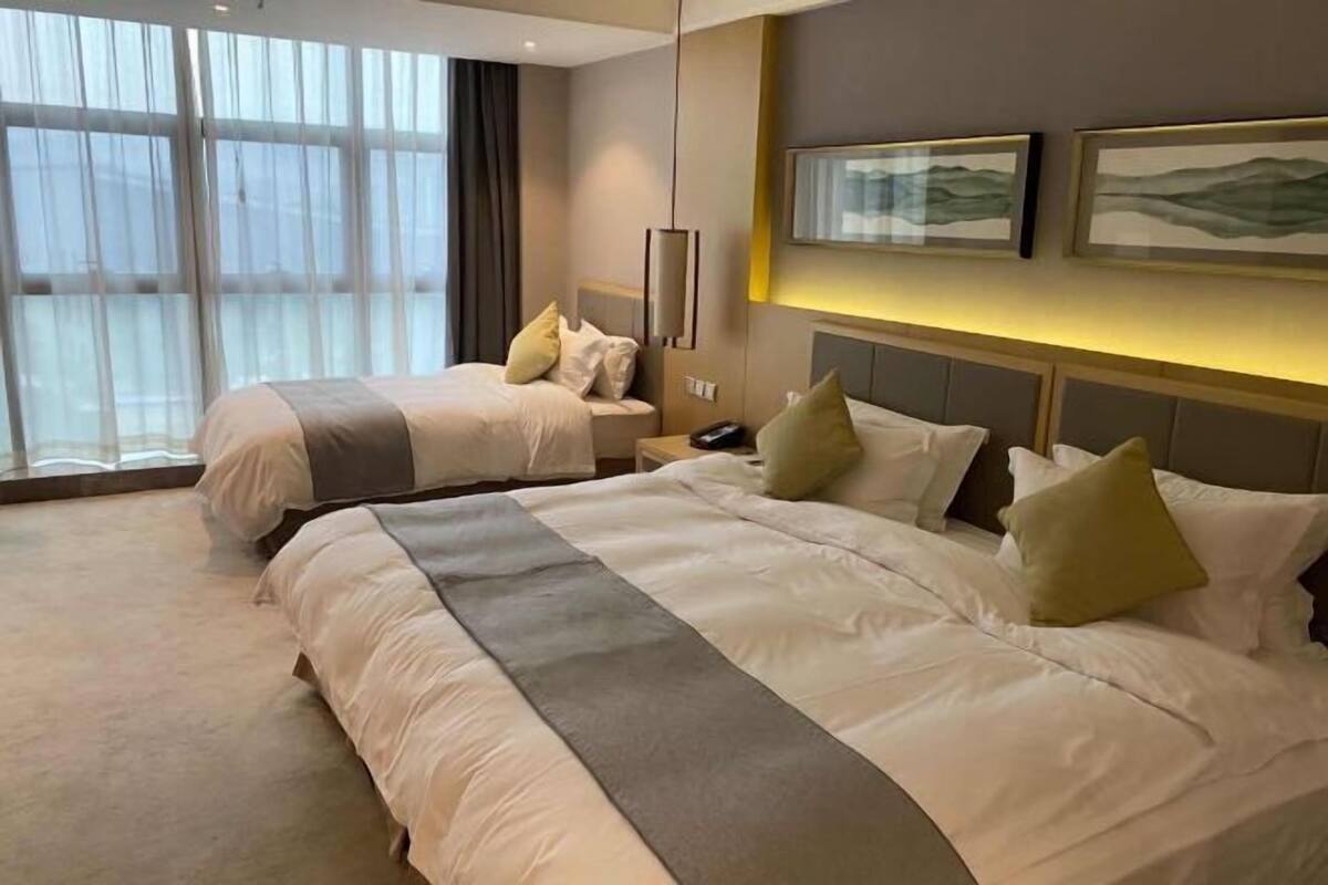A hotel room with two beds, a bedside table with a phone, and large windows with sheer curtains.