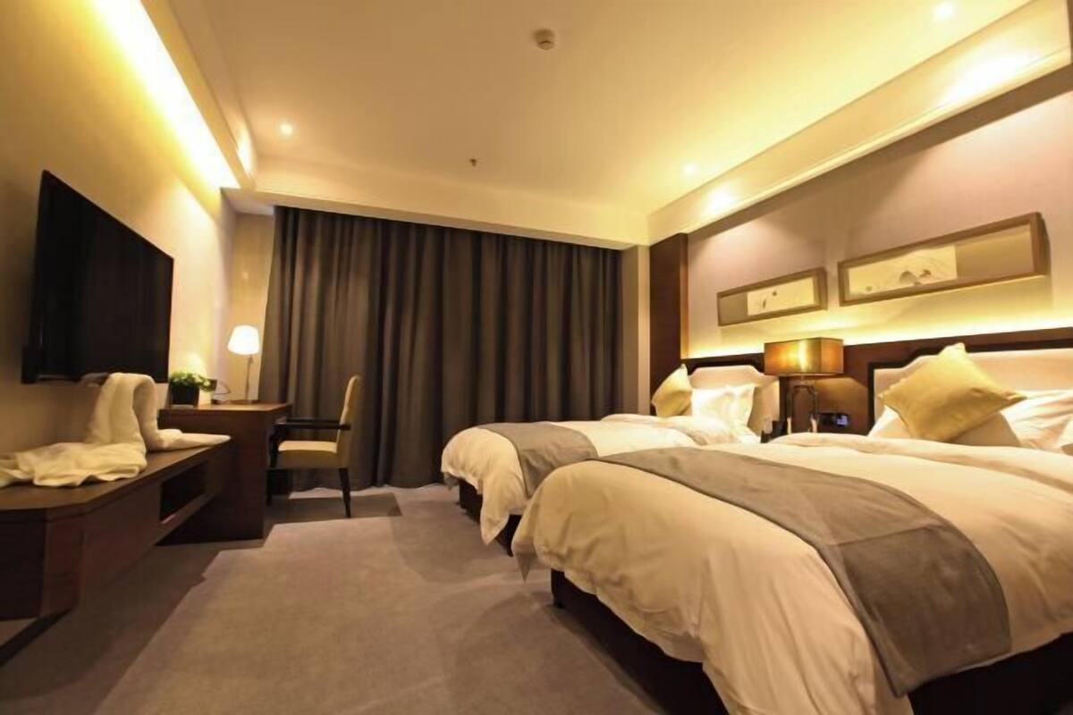 A hotel room with two beds, a desk, a chair, a TV, and a large window with curtains.