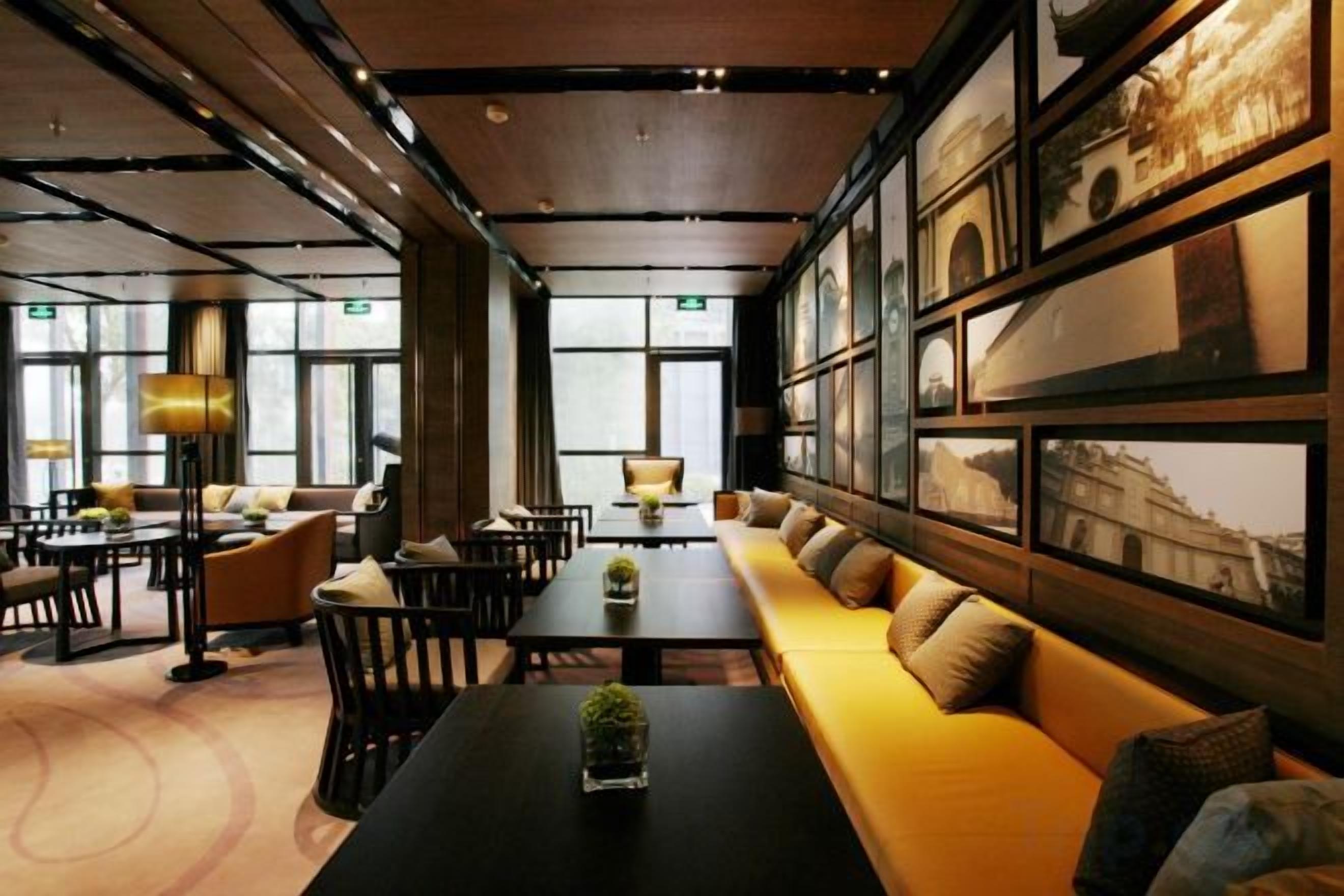 Un vestíbulo de hotel con un sofá largo, mesas de comedor y sillas, ventanas amplias y cuadros en las paredes.