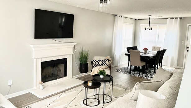 A living room with a fireplace, a television mounted above it, a dining area with a table and chairs, and a large window with curtains.