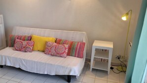 A white sofa with colorful cushions, a white side table, and a floor lamp in a room with white walls and tiled flooring.