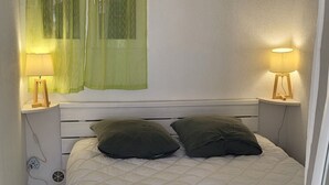 A small, neatly made bed with a white quilt, two green pillows, and a bedside table with a lamp.