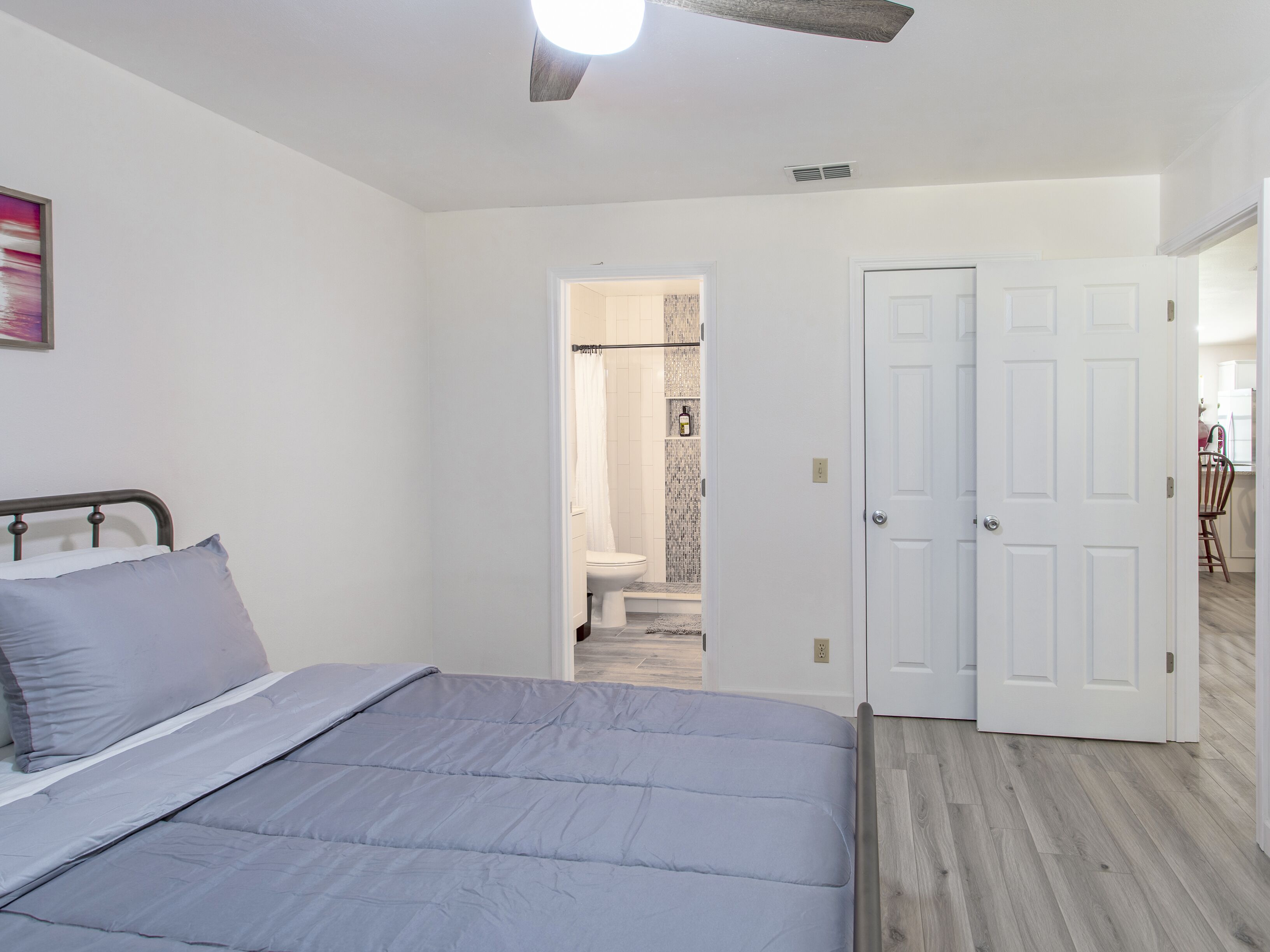 A bedroom with a bed, a door leading to a bathroom, and a view of a kitchen area.