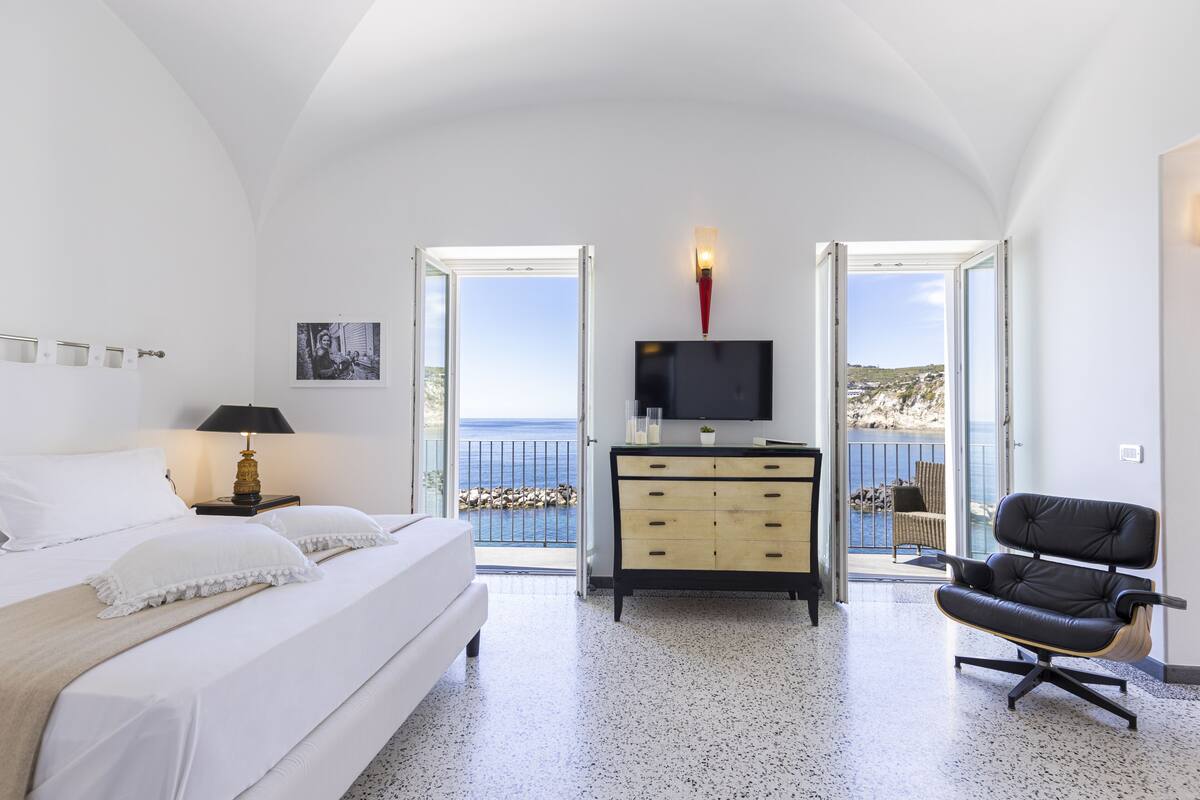 A bedroom with a bed, a TV, a dresser, and a chair. There are two doors leading to a balcony with a view of the sea.