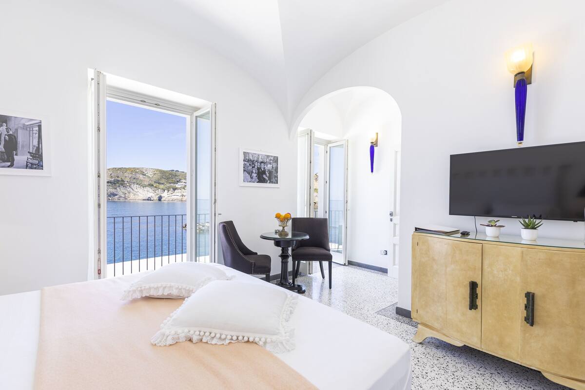 A hotel room with a bed, a TV, a dining table with chairs, and a view of the sea through an open door.