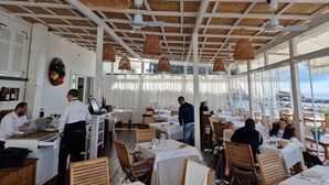 Un restaurant avec des tables blanches et des suspensions en osier, de grandes fenêtres donnant sur la mer et un plafond haut avec des poutres en bois.