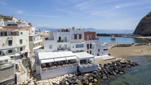 Un bâtiment situé sur la côte, doté de nombreux balcons et d’un bar sur le toit, offrant une vue sur une plage rocheuse et une mer d’un bleu limpide.