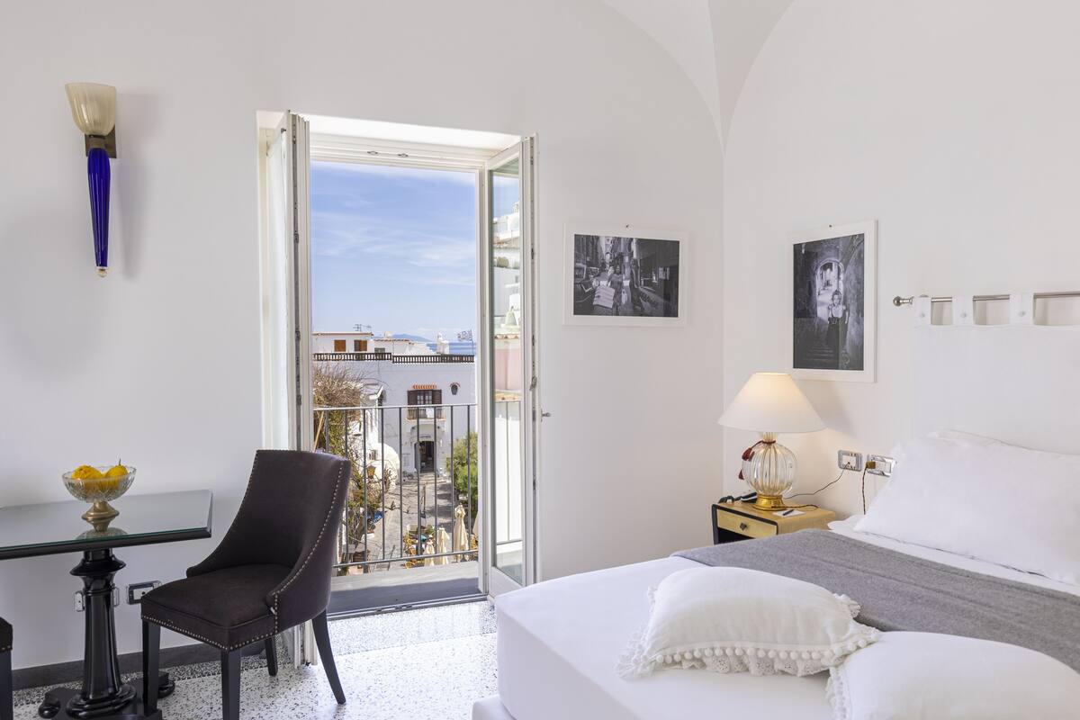 A hotel room with a bed, a chair, a desk, a lamp, and a balcony with a view.