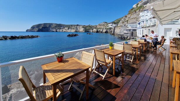 Une terrasse en bois, agrémentée de tables et de chaises, offre une vue sur une zone côtière caractérisée par des falaises rocheuses et une eau d’un bleu limpide.