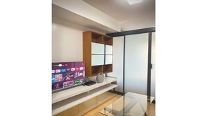 A modern living room with a glass coffee table, a flat-screen TV on a white shelf, and a sliding glass door leading to a balcony. - Convenient 1BR Beside SM Mall (Davao City)