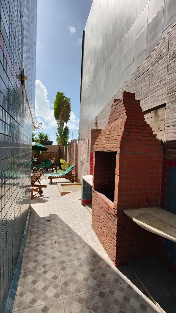 A narrow outdoor corridor with tiled flooring, a brick structure with a small opening, and a green umbrella in the background. - Hotel Pousada Bossa Nova (Maceió)