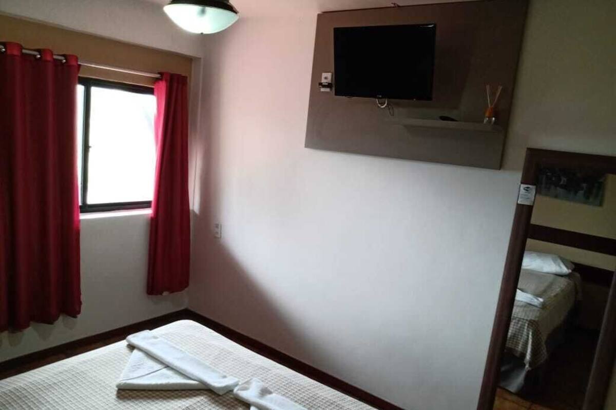 Quarto com cama, televisão na parede e janela com cortinas vermelhas.