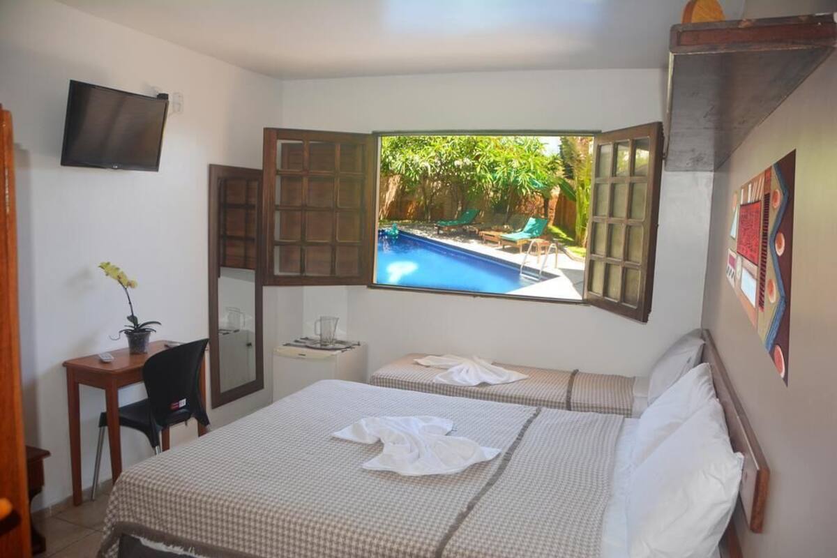Quarto de hotel com cama, escrivaninha, televisão e janela com vista para a área da piscina.