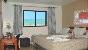 Habitación de hotel con dos camas, una mesita con una planta, una silla y una ventana con vista a la playa.