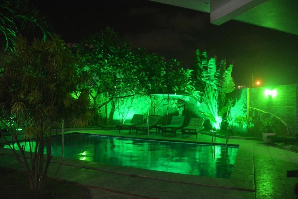 Un área de piscina nocturna con iluminación ambiental verde, sillas de descanso y una piscina cristalina.