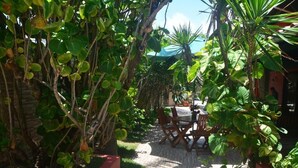 Un sendero en un jardín conduce a una mesa y sillas de madera, rodeado de abundante vegetación y una cerca roja.