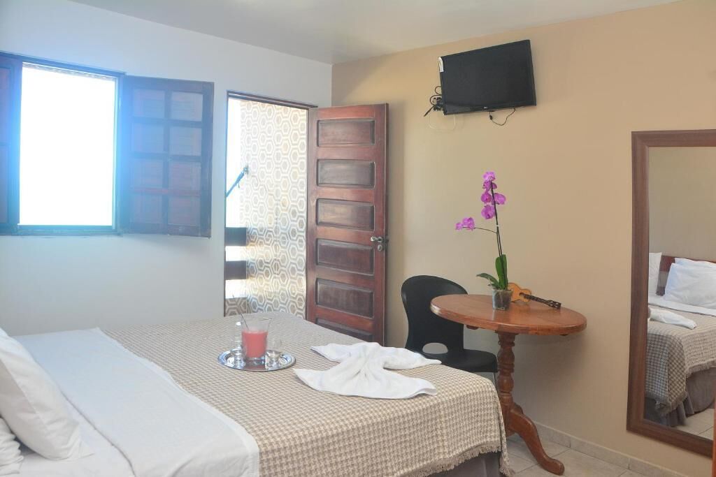 A hotel room with a bed, a small table with a drink and a guitar, a TV on the wall, and a mirror reflecting the room.